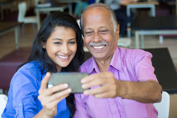 Closeup portrait happy elderly gentleman in pink shirt and lady in blue top taking selfie together, isolated indoors background. Say cheese and smile