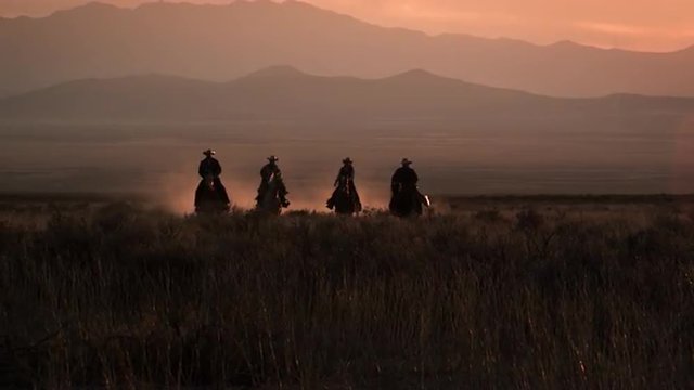 Cowboys galloping against a silhouetted mountain range and orange sunset, Americana.