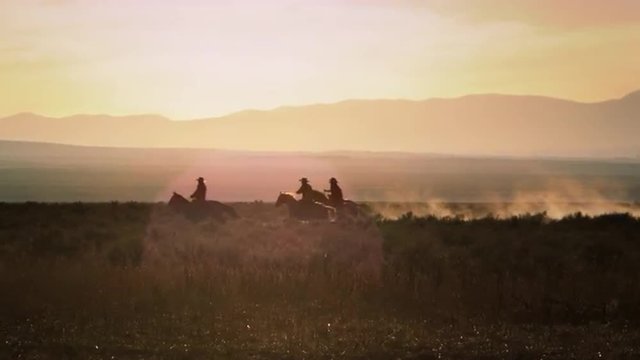Cowboys galloping riding horses accross the dusty western plains against a golden sunset / sunrise, mountains and lens flare.