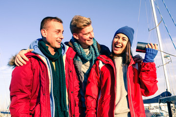 Three young friends having fun before embark on boat at pier - Cheerful people smiling outdoors in sunny  day on winter vacation - Concept of friendship with joyful teenagers playing with a tin