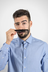 Funny immature guy holding plastic black comb to imitate his fake mustache.  Desaturated portrait over gray studio background with retro vignette. 