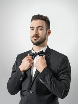 Confident young man in tuxedo with bow tie posing at camera holding suit collar. Desaturated portrait over gray studio background with retro vignette. 