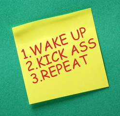 Yellow sticky note with the words Wake Up, Kick Ass and Repeat in red text as a motivational reminder
