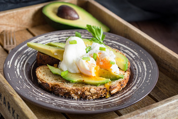 Poached egg and sliced avocado on toasted whole wheat bread on plate. Garnished with scallions and parsley.