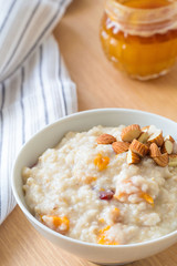 Oatmeal porridge with dried apricots and chopped almonds on bright wooden table. Jar of honey on background. Selective focus. Bright healthy breakfast image