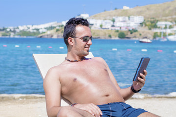 young man laying on a sunbed on the beach relaxing and reading with a tablet