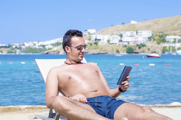 young man laying on a sunbed on the beach relaxing and reading with a tablet