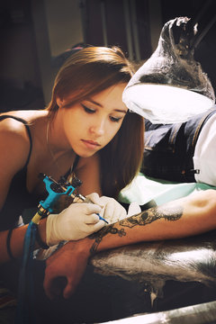 Beautiful young girl tattoo artist portrait during creation tattoo on a man`s hand.