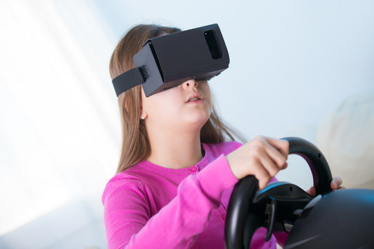 Happy smiling young beautiful young girl holding a gaming computer wheel getting experience using VR-headset glasses of virtual reality at home