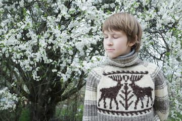 Daydreaming teenage boy with closed eyes wearing knitted wooly reindeer warm sweater outdoors at flowering white fruit tree background