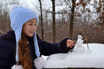 Юная девочка в зимнем заснеженном лесу слепила маленького забавного снеговика.