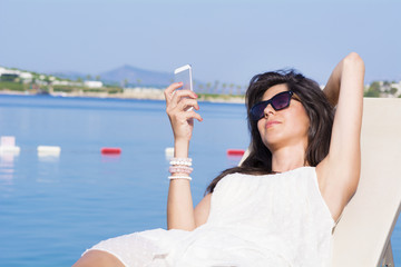 Portrait of young beautiful woman with white dress  laying on a sunbed and sending messages on a sea background.Summer vacation