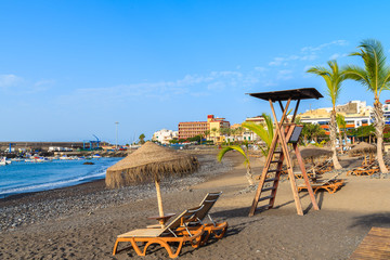 Palm trees and sunbeds on tropical beach in San Juan town on coast of Tenerife, Canary Islands,...