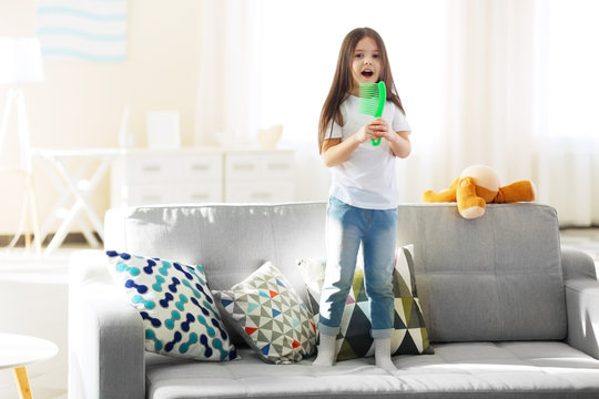 Beautiful little girl singing in a green hairbrush on sofa in the room