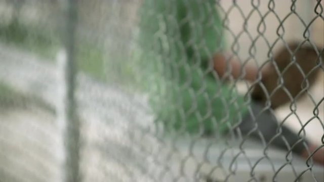 Slow motion rack focus of boy sitting in dugout behind chain link fence.