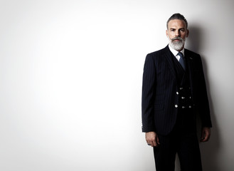 Portrait of bearded gentleman wearing trendy suit and stands against the empty white wall.