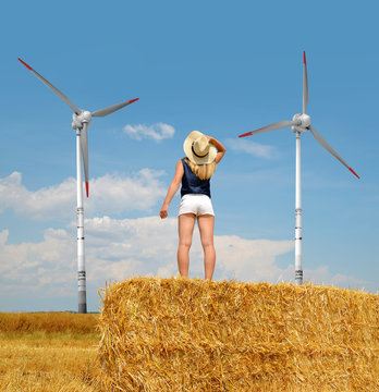 Woman with hat standing on a bale of straw. In the background wind turbines.