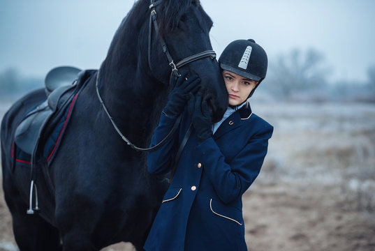 A Tall Girl With Long Blond Hair In A Jockey Outfit With A Beautiful Black Horse In An Empty Snow-covered Field In Winter