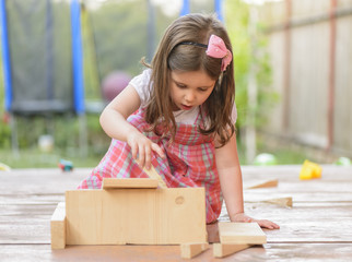 Portrait of beautiful little girl playing outside with wooden blocks, being inventive building a house for her toys