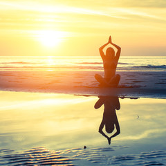 Yoga woman sitting on the beach with reflection in water, during sunset.