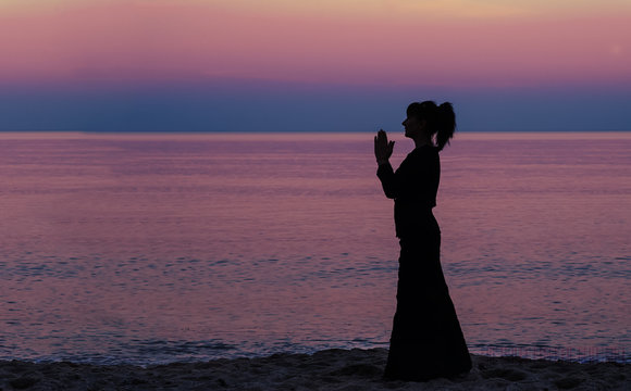 Silhouette of a woman standing on beach with hands in prayer position 