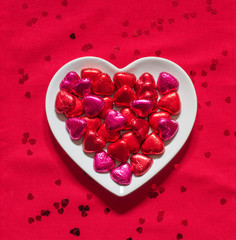 A red, heart shaped plate full of Valentine's Day chocolates on a red tablecloth.