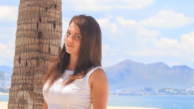 girl with big bust in white smiles by palm trunk on beach