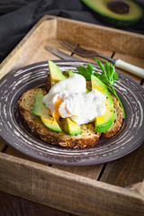 Poached egg and avocado on toasted whole wheat bread with seeds on a plate in vintage wooden tray. Garnished with scallions and parsley.