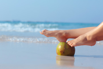 Female feet propped on coconut on the beach, blue sea background