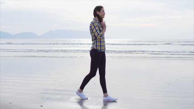 Slow motion video of young woman walking on beach. Full length side view of beautiful female in casual wear. Tracking shot of woman enjoying nature. Waves are rushing towards shore in background.