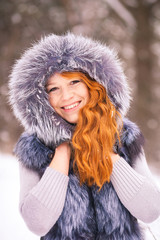 Winter girl. Portrait of beautiful young girl dressed in fur coat. Winter background. Beauty woman having fun outside on winter snowy day. Close up of female smiling face with gorgeous long red hair