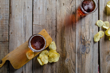 Potato chips scattered on a table and two glasses of dark amber beer against a wooden rustic cuting board on textured background, top view.