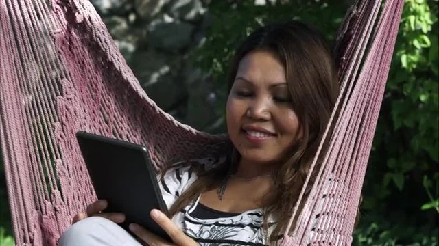 Handheld shot of Asian woman using a tablet and glancing at the camera while sitting on a hammock