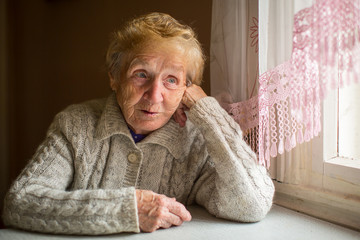 An elderly woman sits in the house near the window.