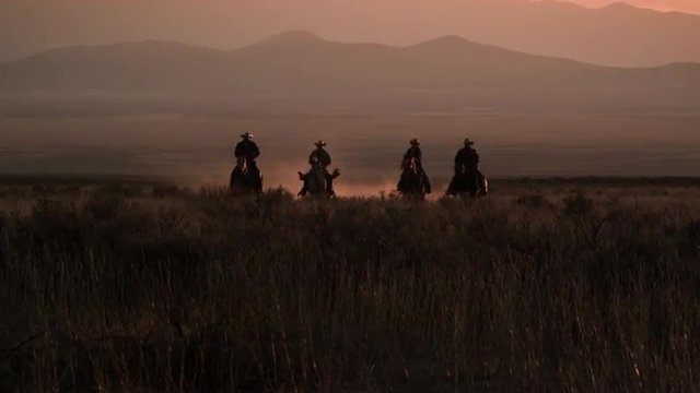Slow motion shot of cowboys galloping in distance.