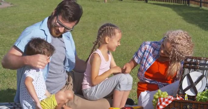 Young parents kissing and hugging their cute little children during family picnic on green lawn 
