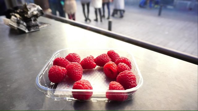 Human hand picking red raspberry in the plastic box with people walking background 