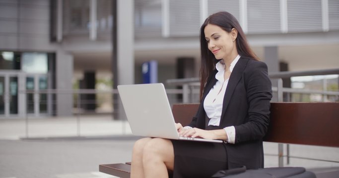 Cute business woman sitting alone on bench in front of office building working on laptop computer