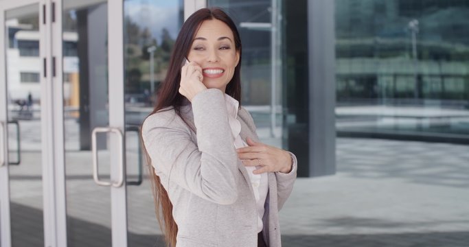 Excited young brunette businesswoman with big smile on phone near glass door outside with hand on chest