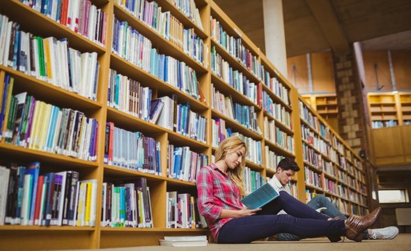 Smiling classmates reading book while leaning on bookshelves