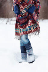 The figure of a young woman in bright ethnic scarf with purple, white and blue pattern, blue jeans and soft boots. She stands in the snow.