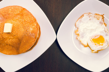 Plates touching on dark surface, one with fried egg, other has pancakes, seen from above, elegant presentation