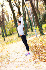 Young woman with headphones preparing for a jogging in autumn park, she is stretching her arms.