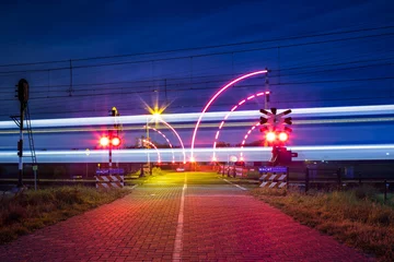Fototapeten Bewaakte spoorwegovergang met brandende lichten in de nacht en voorbijrijdende trein © Leandervasse