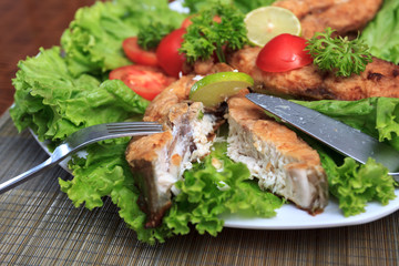 A piece of fried fish fillett served on blue glass table with lettuce, lime, tomato and herbs