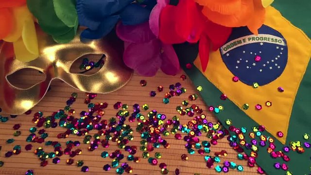 Sparkly carnival sequins falling on top of golden carnival mask with colorful lei and Brazilian flag on textured wood background