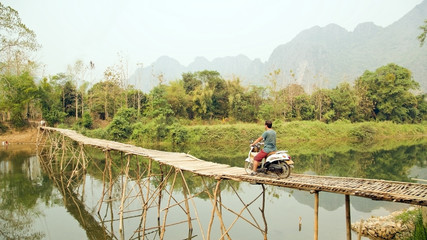 Fototapeta na wymiar Cheerful Tourist crossing bamboo bridge motorbike, limestone view, laos