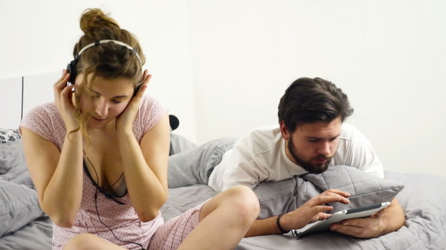 woman listening to music in headphones, man using tablet in bed