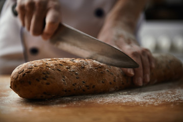 Close-up of baker hands cutting fresh bread from oven. Female cook in uniform preparing meal in restaurant kitchen. 