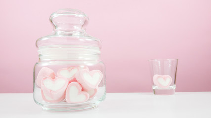 The lovely pink heart marshmallows in small glass jar and shot glass on white table for Valentine's day.